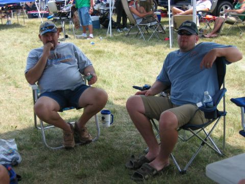 1985 Ford Crown Victoria LTD - Two men relax in camping chairs on a grassy field, with tents and other people visible in the background.