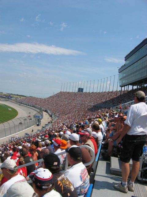 1985 Ford Crown Victoria LTD - Spectators fill the grandstands at a race track, with cars visible on the track in the distance.