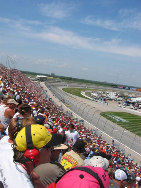 1985 Ford Crown Victoria LTD - A wide view of a race track and the surrounding grandstands filled with spectators on a sunny day.