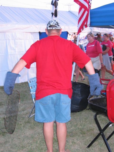 1985 Ford Crown Victoria LTD - A man prepares for a barbecue at an outdoor event, with tents and other people in the background.