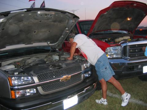 1985 Ford Crown Victoria LTD - A man examines the engine of a silver pickup truck, with another red truck visible behind it.