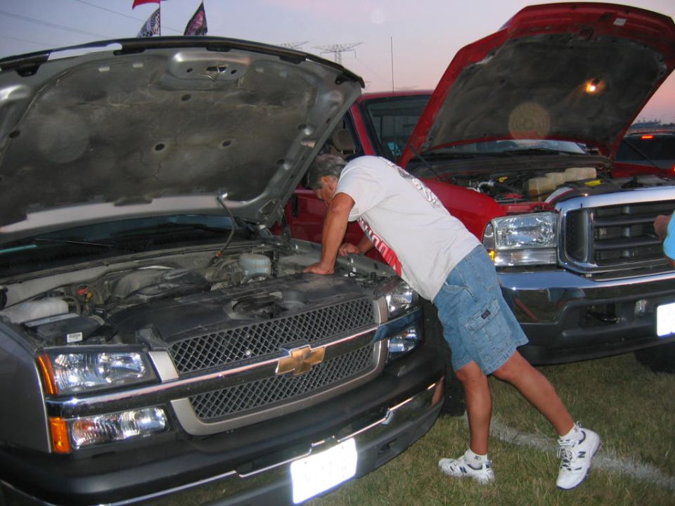 Man inspects engine of silver pickup truck with hood open, red truck behind