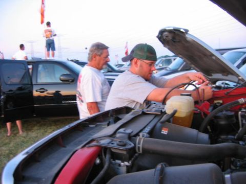 1985 Ford Crown Victoria LTD - Two men are focused on the engine of a vehicle while others stand around at an outdoor event.
