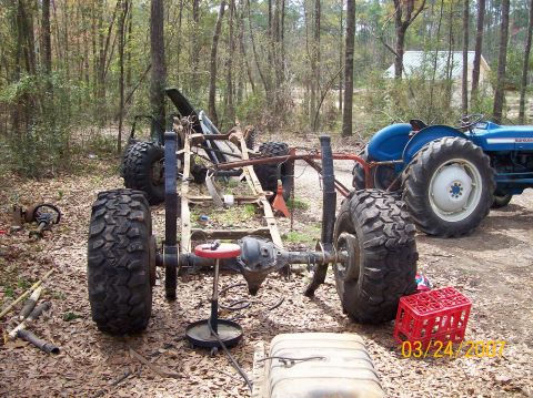 1978 Ford Bronco - A vehicle frame sits on large tires next to a blue tractor, with a red crate and fuel tank nearby.