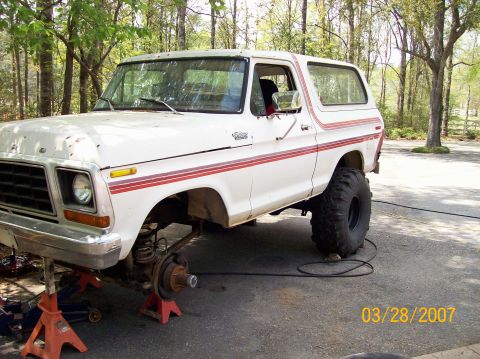 1978 Ford Bronco - A white 1978 Ford Bronco is shown on jack stands with its front wheel removed, indicating it is undergoing maintenance.