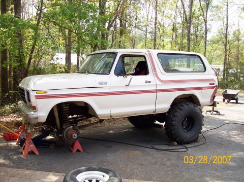 1978 Ford Bronco - A white 1978 Ford Bronco with red side stripes is elevated on jack stands, showing its suspension and large off-road tires.
