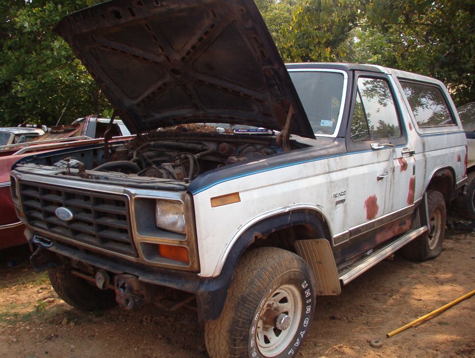 White and blue Ford Bronco with hood up in a salvage yard