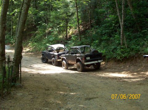 1978 Ford Bronco - Two off-road vehicles navigate a wooded trail on a sunny day.