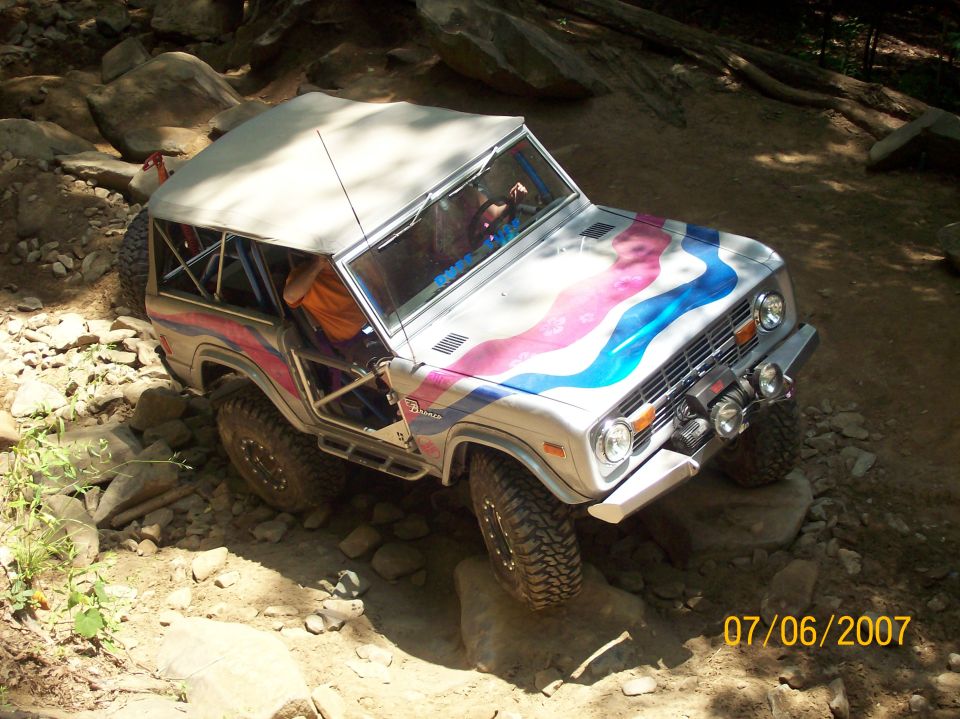 Silver Ford Bronco with pink and blue stripes and soft top climbing rocks on a wooded trail