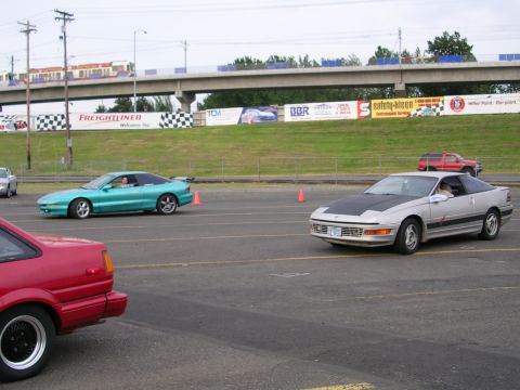 1996 Ford Probe - A silver Ford Probe with a black hood drives on a track, with other vehicles and banners in the background.