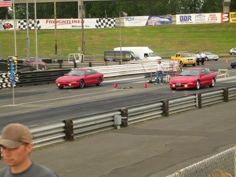 1996 Ford Probe - Two red 1996 Ford Probes compete on a drag strip, with a yellow truck and other vehicles visible in the background.