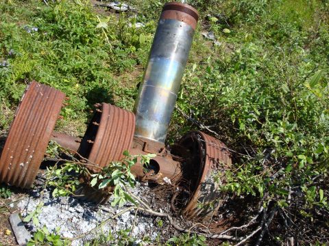 2000 Ford E350 - Discarded vehicle parts, including a large exhaust pipe and two rusty wheel rims, are overgrown with vegetation.