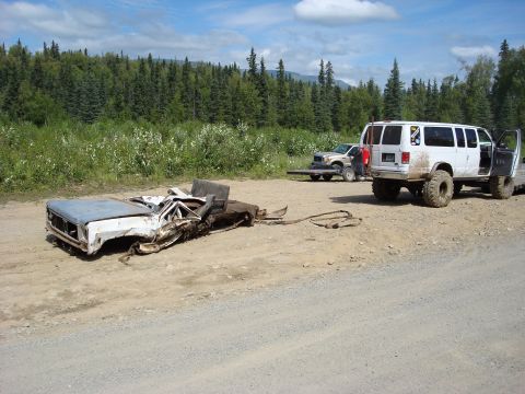 2000 Ford E350 - A severely damaged truck chassis sits on a trailer, with a lifted van nearby in a rural setting.