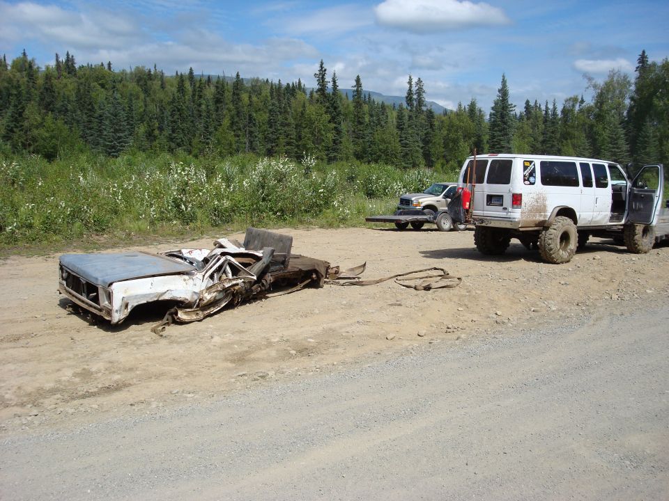 Wrecked truck chassis on a flatbed trailer, with a lifted white van parked nearby. Trees and a grassy field in the background