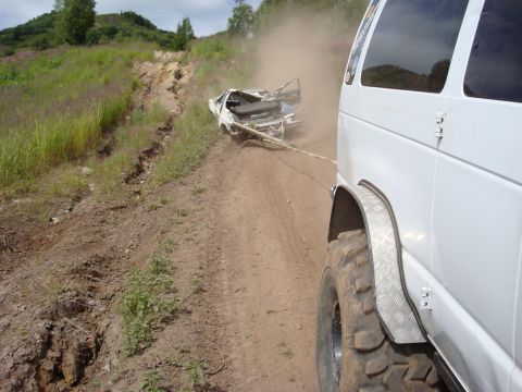 2000 Ford E350 - A white van tows a heavily damaged car up a dusty, off-road incline.