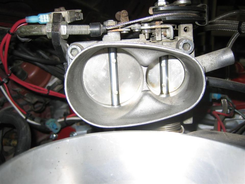 Aluminum twin-bore throttle body with linkage and red plug wires, under the hood.