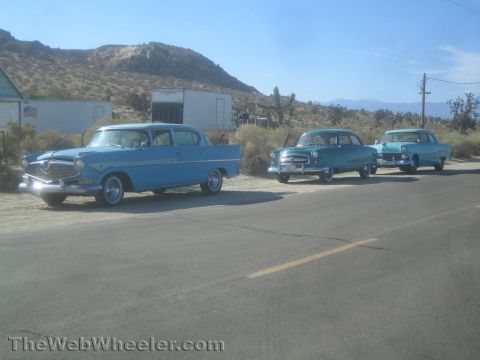 1990 Ford Bronco - Three blue and teal 1950s sedans parked along a sunlit desert road with mountains in the background.