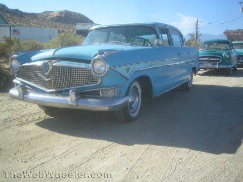 1990 Ford Bronco - Front three-quarter view of a light blue 1950s Hudson sedan with chrome grille parked on a desert road, another car behind.