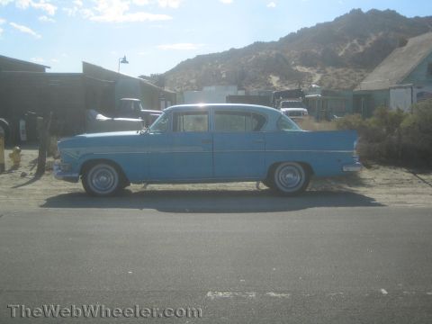 1990 Ford Bronco - Side profile of a light blue 1950s four-door sedan parked along a desert road with mountains behind.