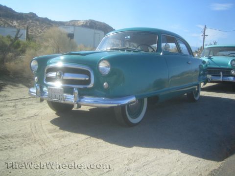 1990 Ford Bronco - Front three-quarter view of a teal 1950s Nash compact car with chrome grille and whitewall tires parked on sand.