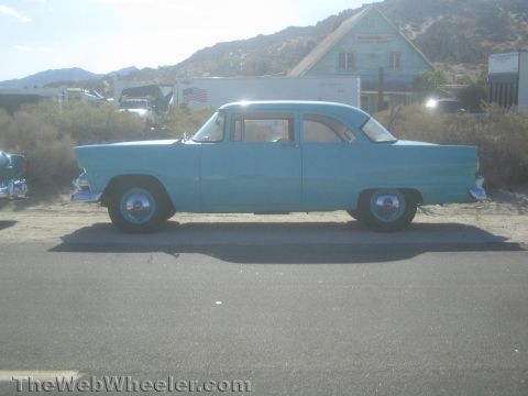 1990 Ford Bronco - light blue 1950s four-door sedan parked alongside a desert road, with mountains and a building in the background.
