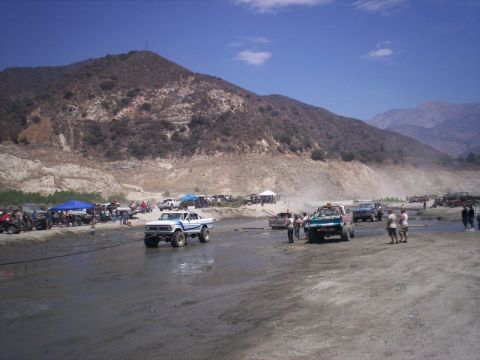 1985 Ford Bronco - Several modified trucks are gathered in a dusty, off-road environment with spectators present.