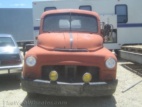 1990 Ford Bronco - front of a weathered orange vintage pickup truck with round headlights and fog lamps, parked in a dirt lot beside trailers.