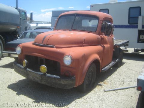 1990 Ford Bronco - weathered orange vintage flatbed pickup truck with round headlights and yellow fog lamps, parked among trailers in a gravel yard.