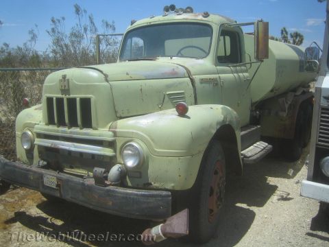 1990 Ford Bronco - pale green vintage International Harvester truck fitted with a rear tank, parked on a dirt lot under a blue sky.