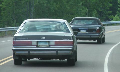 1991 Ford LTD Crown Victoria - A silver Mercury Grand Marquis LS and a dark sedan drive on a two-lane road bordered by trees.
