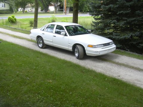 1997 Ford Crown Victoria - A white sedan is parked on a gravel driveway, with trees and grass in the background.