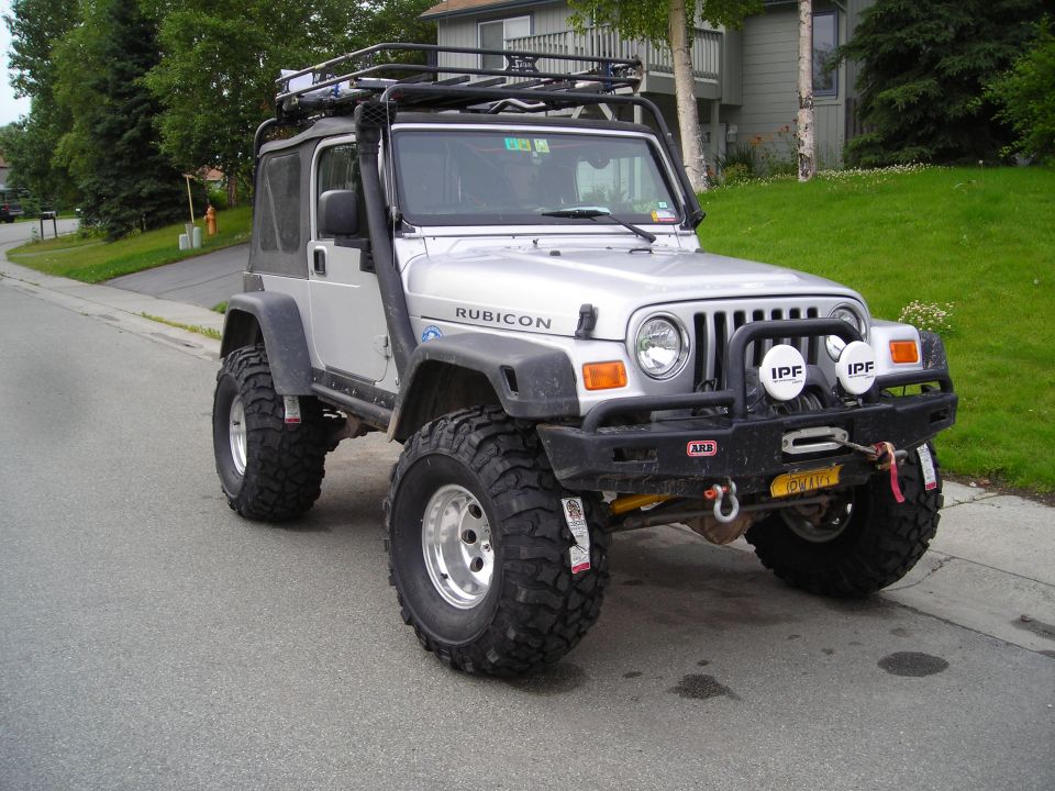 Silver Jeep Wrangler Rubicon with roof rack and snorkel. It has large off-road tires and a winch on the front bumper