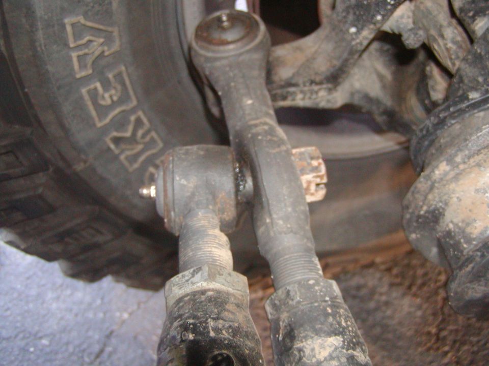 Close-up of vehicle steering linkage components next to a tire with 'KEW' visible on the tread. Supermotors.net logo in lower right