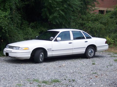 1994 Ford Crown Victoria - A white 1994 Ford Crown Victoria is parked on a gravel lot with trees and a building in the background.