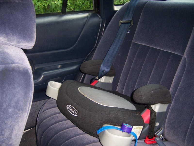 Child booster seat on dark blue velour rear car seat, with cup holders and seatbelt visible