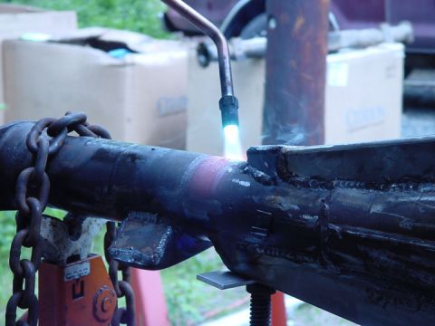 1979 Ford Bronco - A metal axle housing is being heated with a torch, with chains attached and supported by a jack.