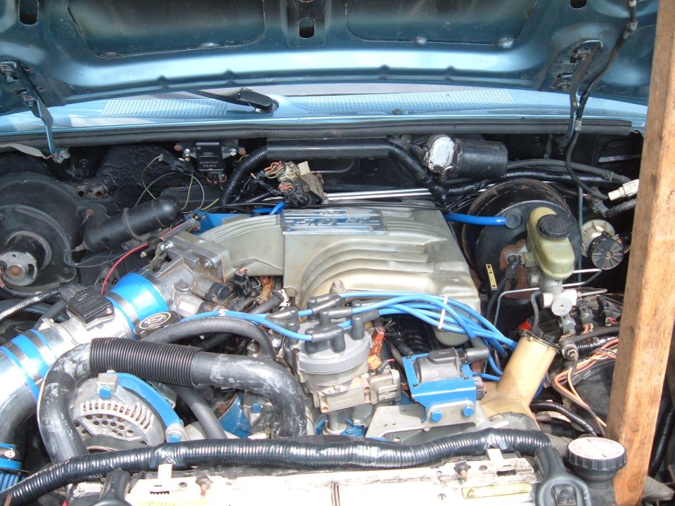 Engine bay with a V8 engine, blue hoses, and silver intake manifold. Some wiring and engine components are visible