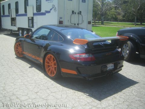 1990 Ford Bronco - black Porsche 911 GT3 RS with orange wheels, side stripe and rear wing, parked on gravel near a trailer.
