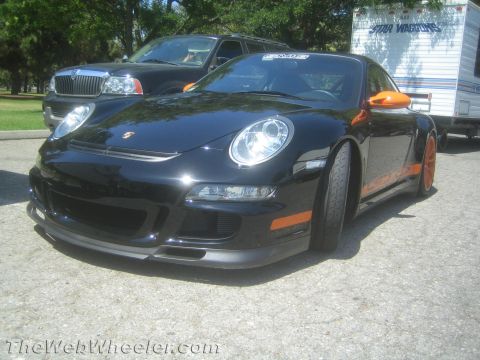 1990 Ford Bronco - black Porsche 911 GT3 RS with orange side stripe and bright headlights, parked on gravel beside other cars and a trailer.