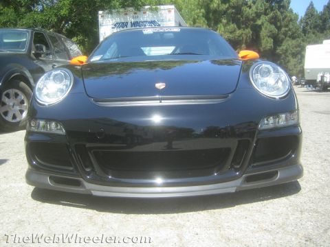 1990 Ford Bronco - front of a black Porsche 911 GT3 RS showing round headlights, low bumper splitter and an orange tow strap, parked on gravel.