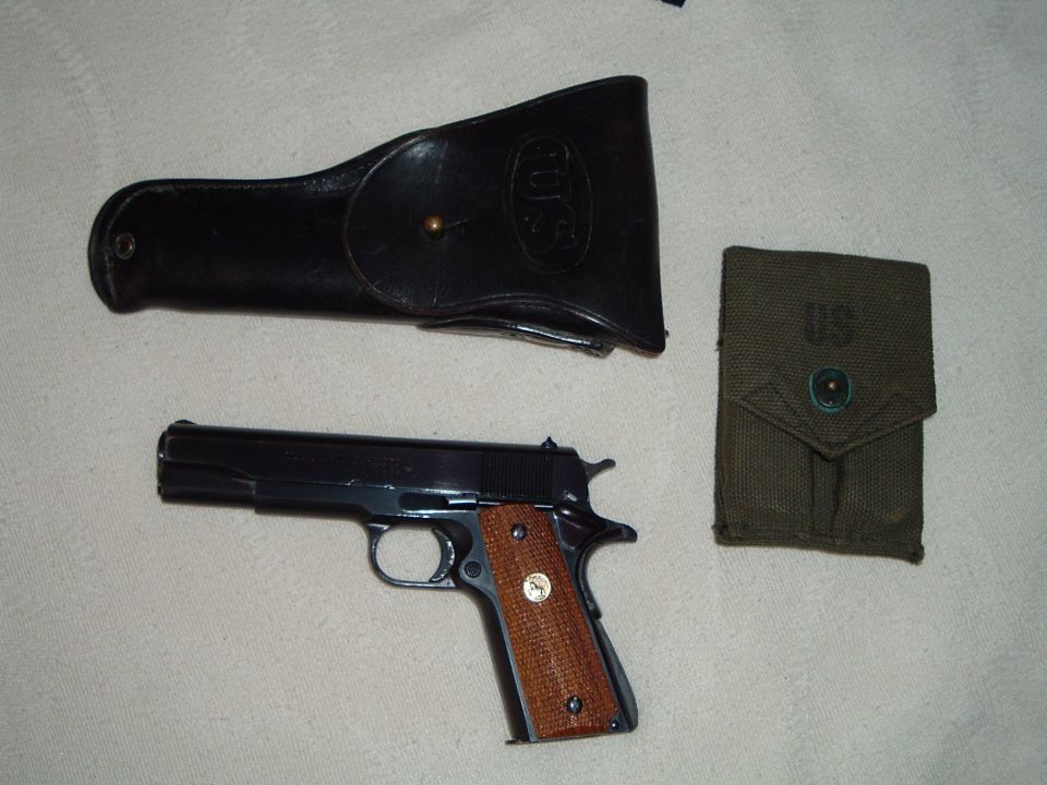 Colt M1911 pistol, black, with brown checkered grips, next to a black leather US marked holster and a green canvas US marked pouch