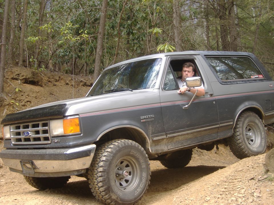 Gray two-tone Ford Bronco with big mud tires on a dirt trail, driver leaning out the window