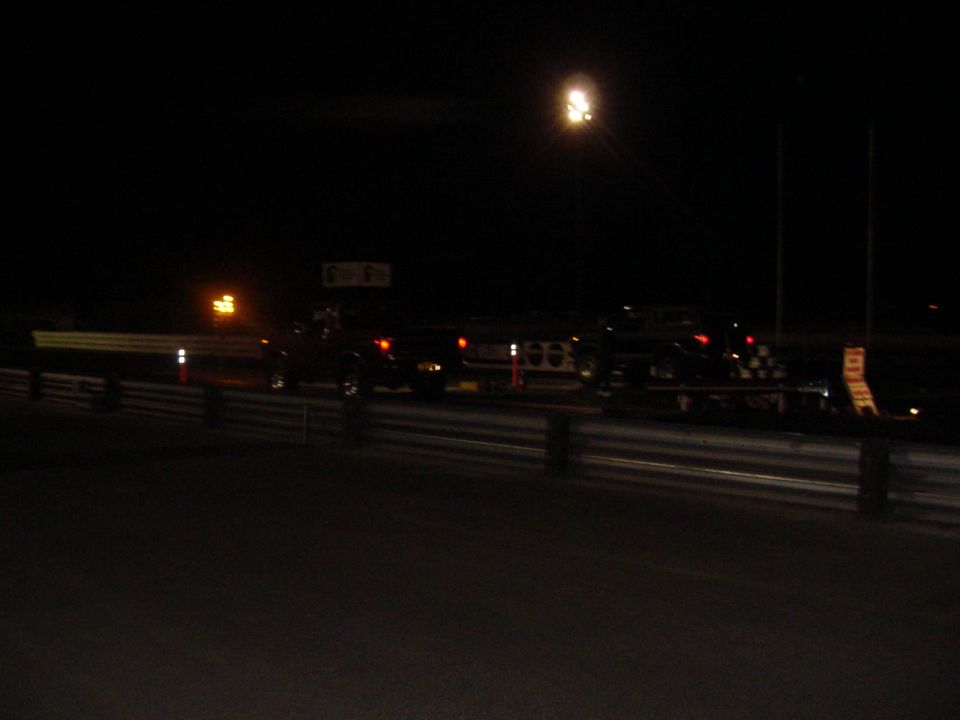 Dark nighttime scene at a drag strip with vehicles and a tow light under a pole lamp