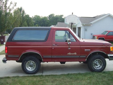 1987 Ford Bronco - A red 1987 Ford Bronco 4x4 SUV is parked on a driveway with a black removable top and chrome wheels.