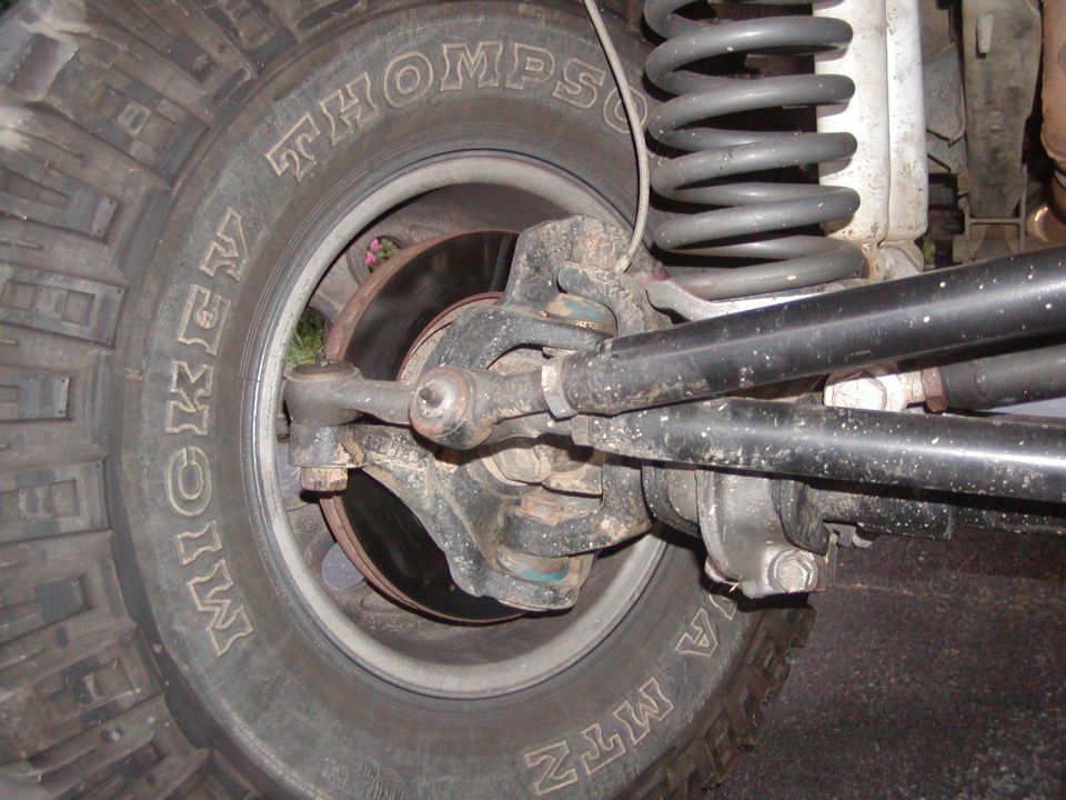 Close-up of a vehicle's front wheel, suspension, and steering linkage. Mickey Thompson tires are visible