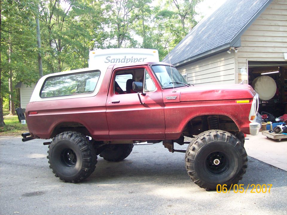 Lifted red Ford Bronco with white hardtop on huge tires parked by a garage and camper