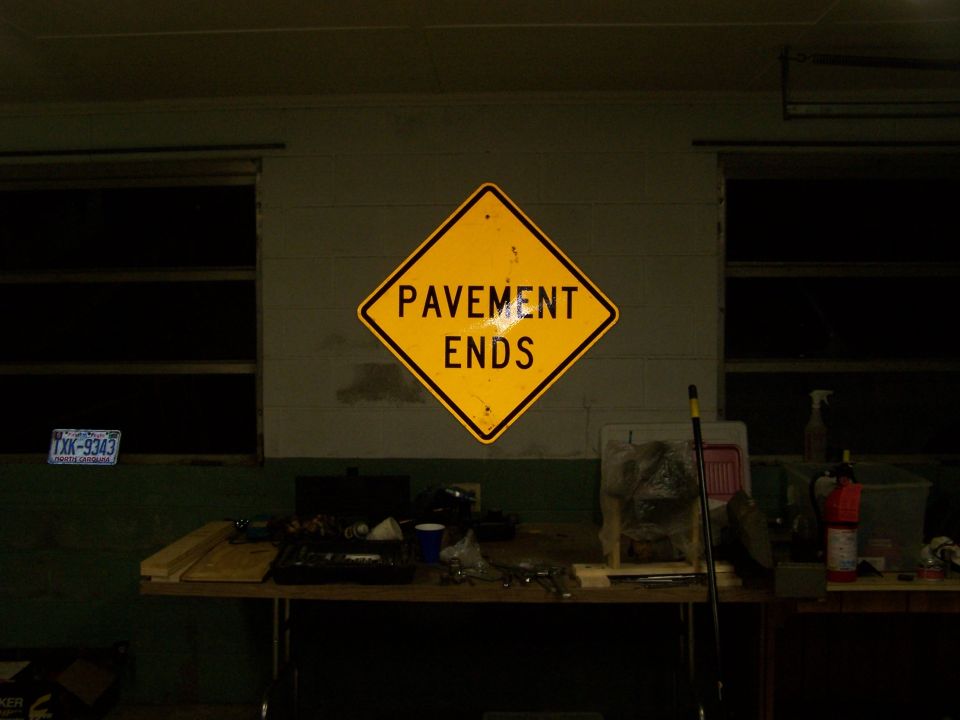 Yellow diamond sign reading 'PAVEMENT ENDS' hangs on a wall above a table with tools