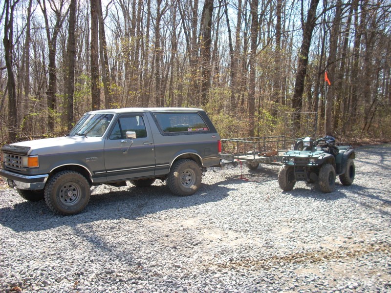 Gray lifted Ford Bronco towing a trailer with a green Honda ATV on gravel by woods.