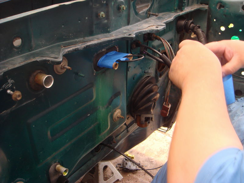 Car firewall with green paint, showing wiring harness and blue tape on a grommet. A person's hands are visible working on the wires