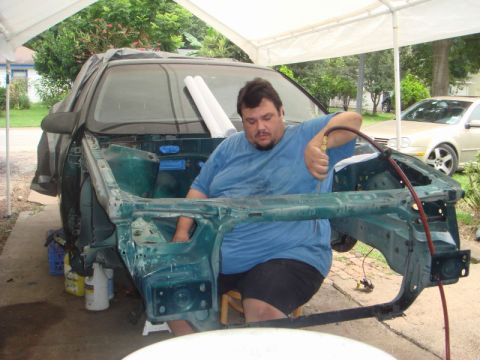 1993 Ford Taurus - A man works on the green frame of a disassembled vehicle under a canopy, with another car visible in the background.